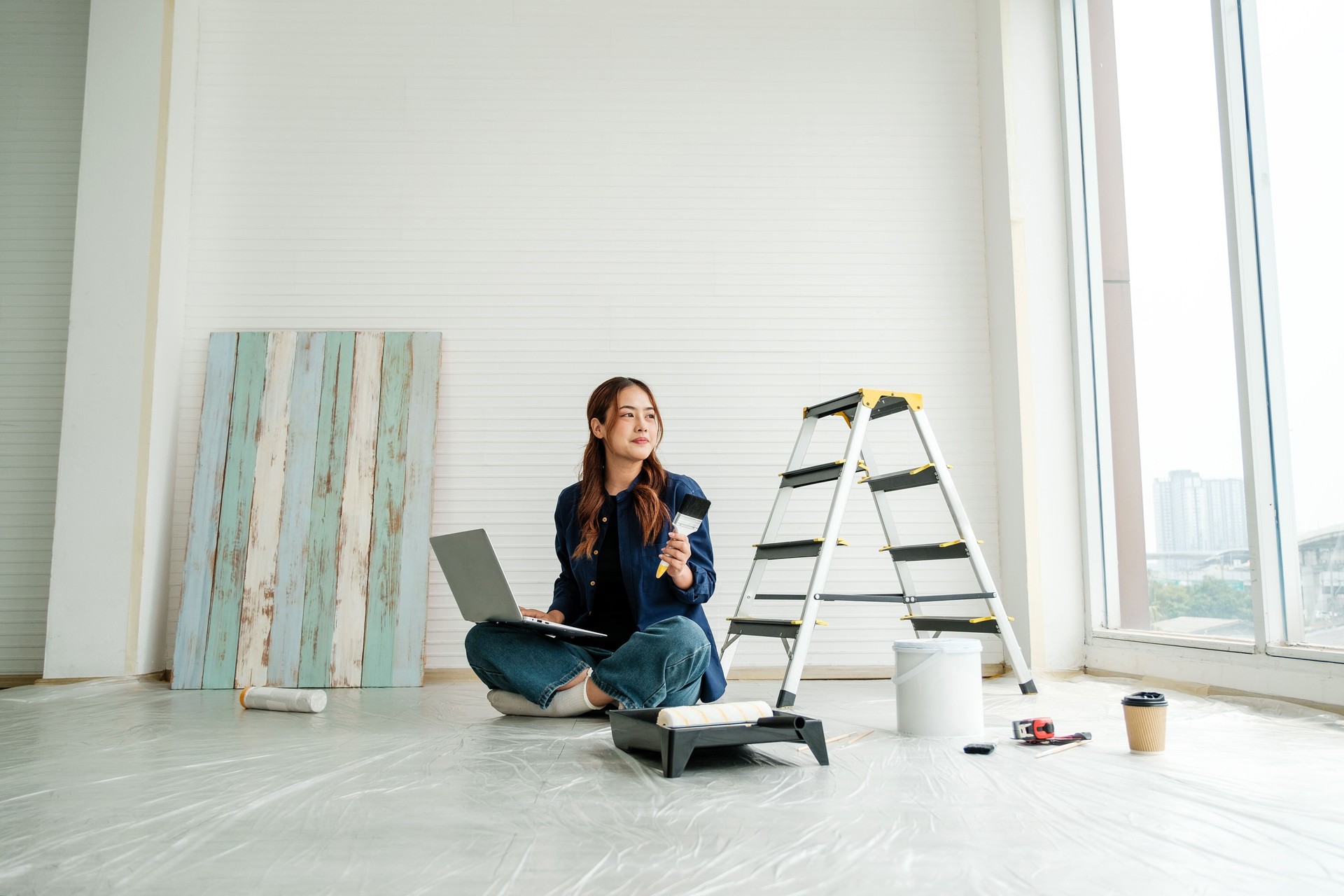 Young woman doing a DIY home renovation project, holding a paintbrush and using a laptop. Creative interior design and home improvement concept with tools, ladder, and coffee in a bright space.